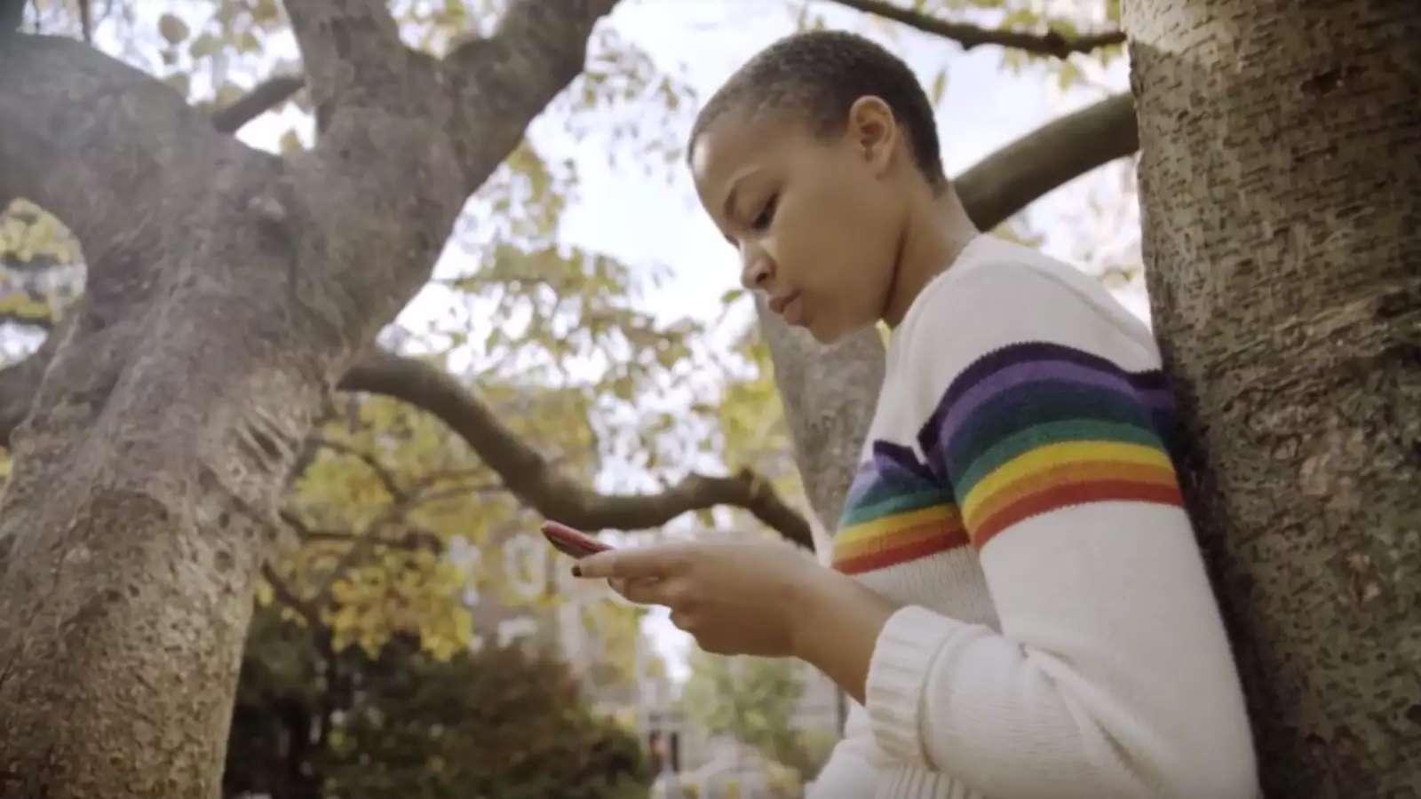 Black woman student leaning against a tree, looking at her phone